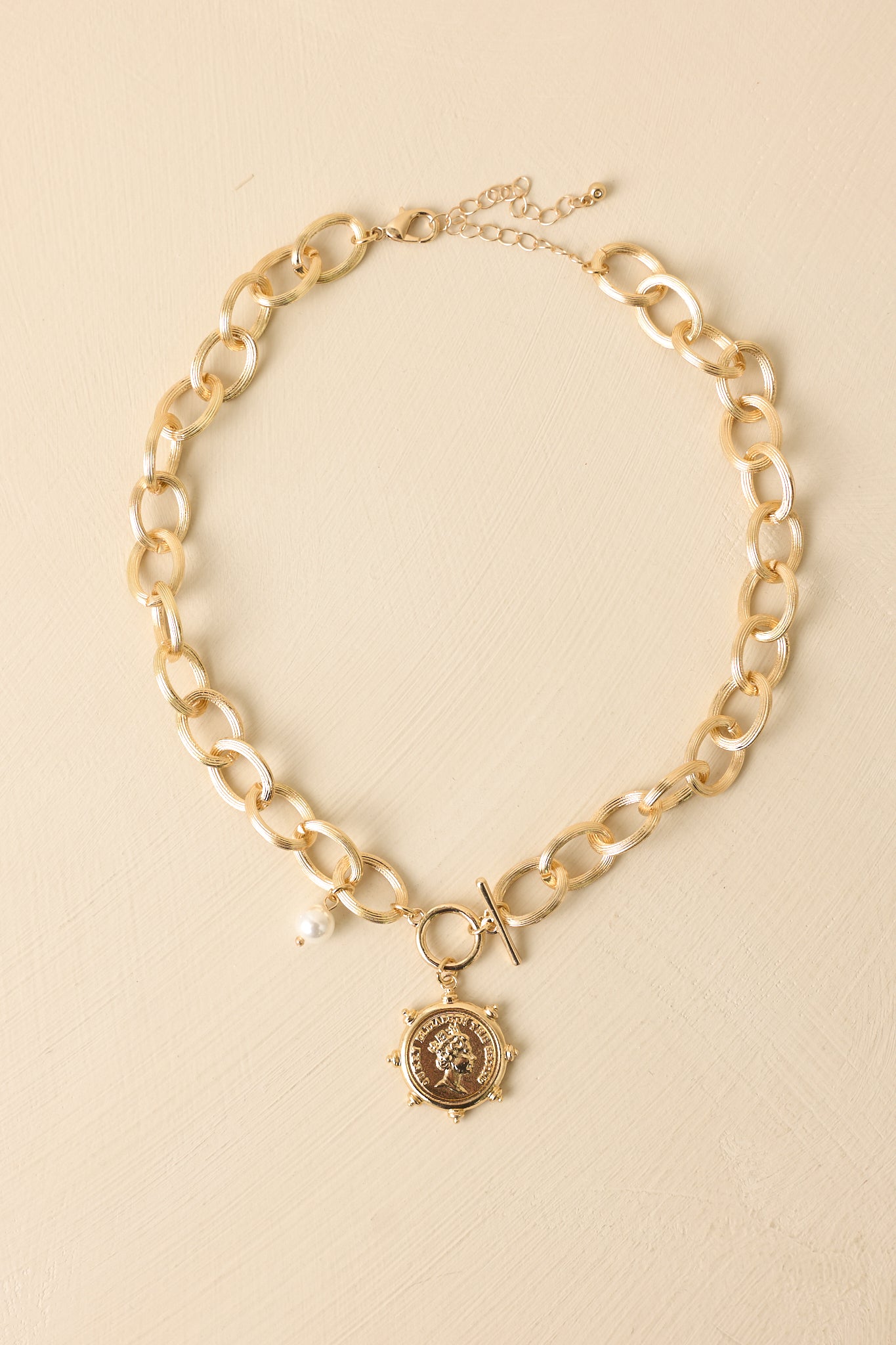 Gold statement chain necklace displayed on a beige background showcasing oversized links and coin-style pendant charm.