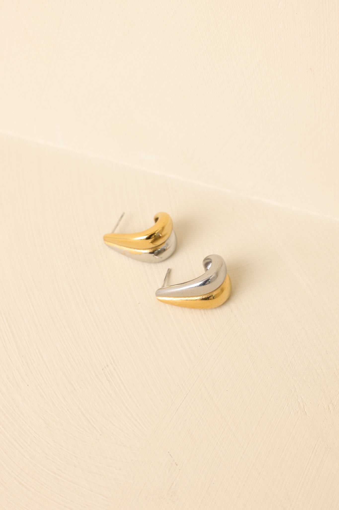 Angled view of mixed-metal sculptural earrings on beige backdrop highlighting shine and dimensional form.