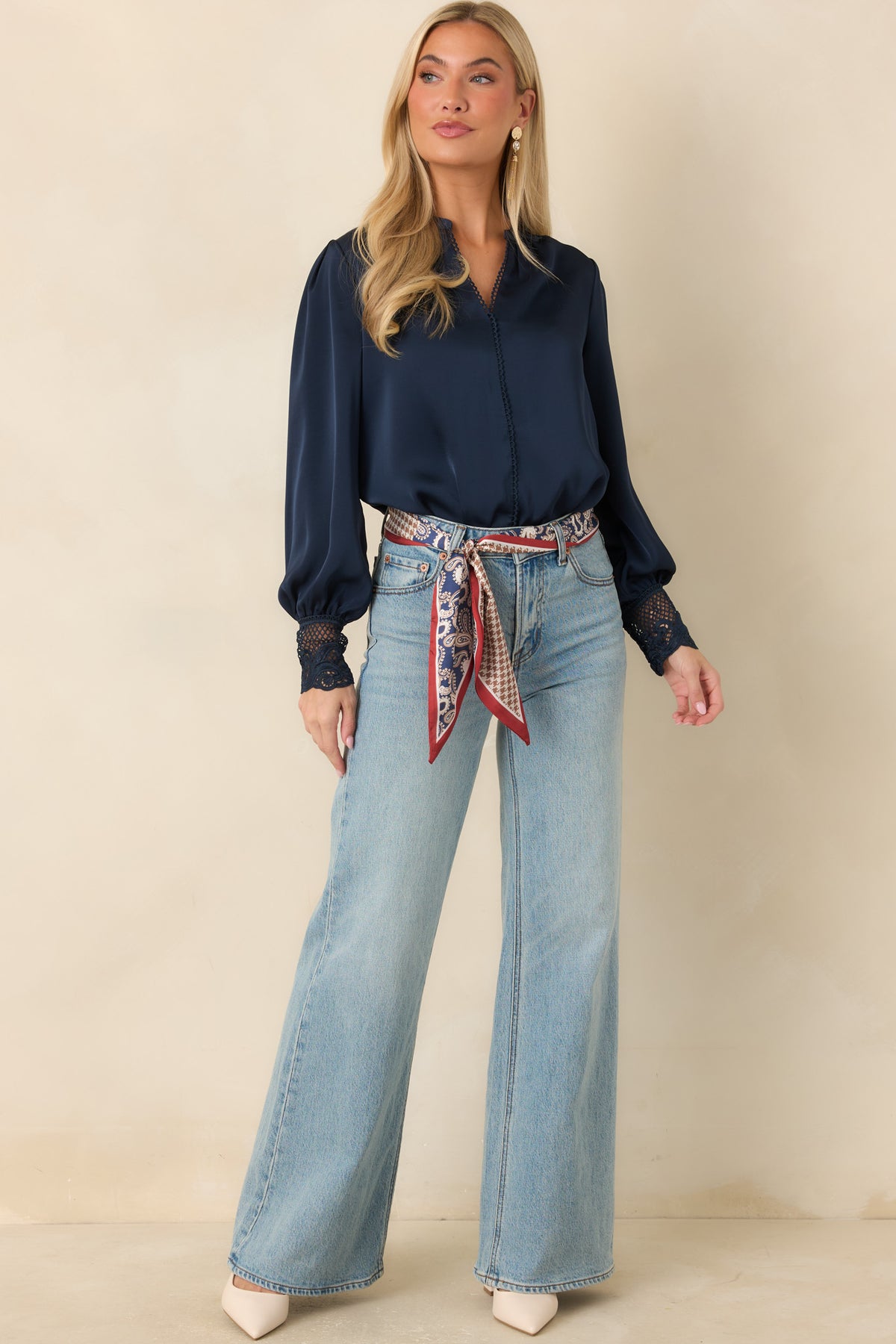 A woman modeling a navy blue long-sleeve blouse with sheer lace cuffs, paired with light-wash wide-leg jeans and a red patterned silk tie belt, against a solid beige studio background.
