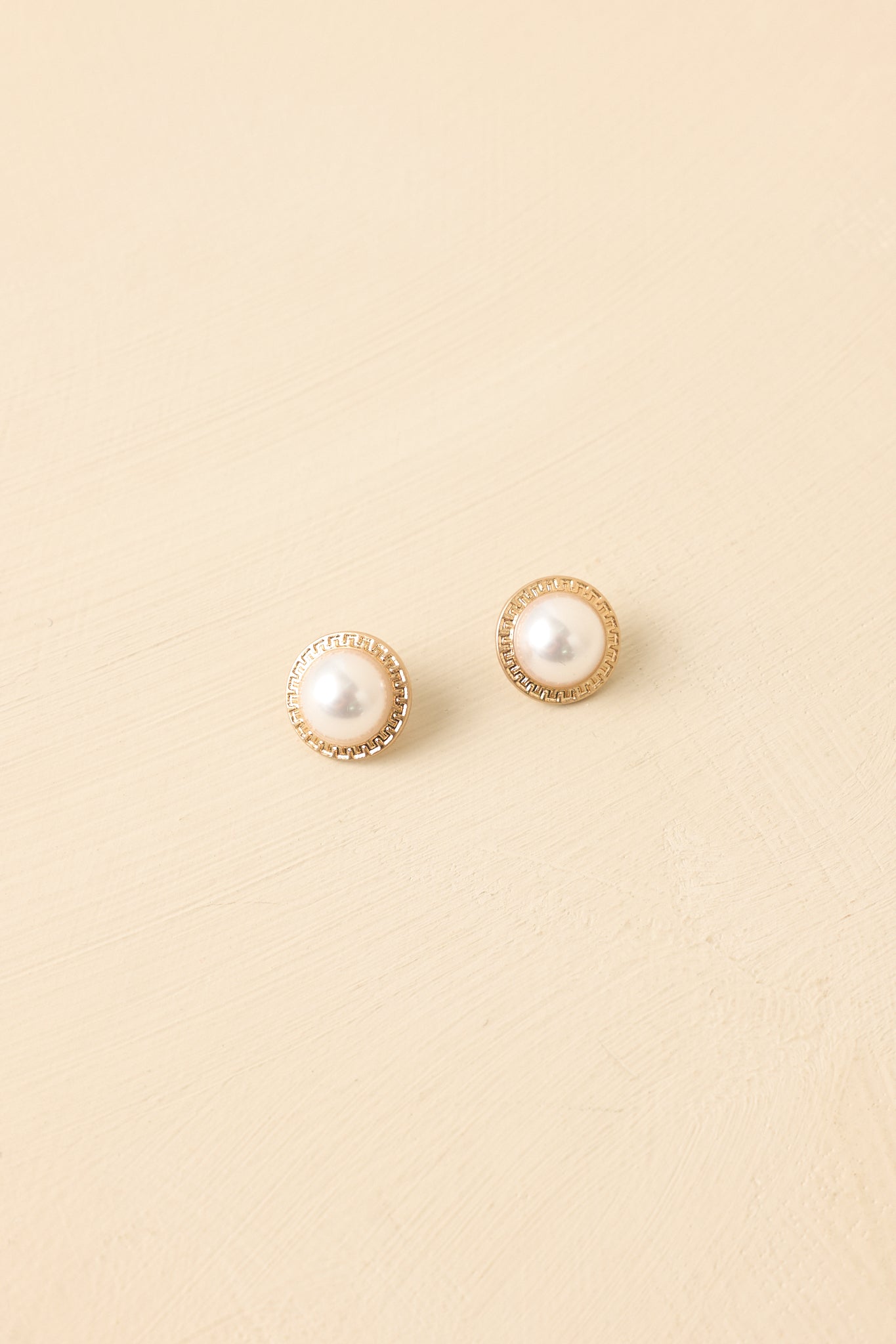 Front view of pearl stud earrings on beige background showing gold bezel framing and smooth pearl surface.