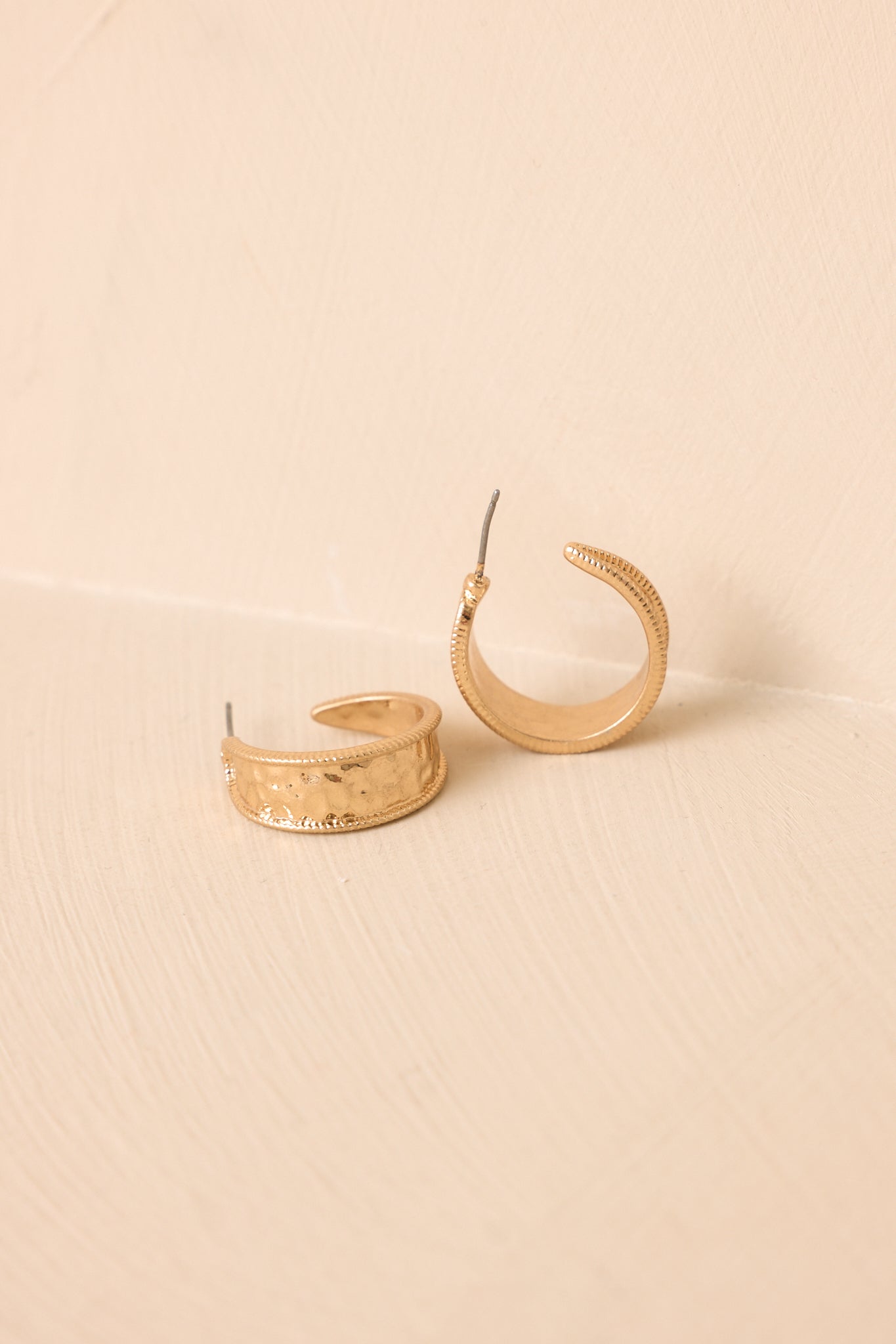 Pair of gold hammered hoop earrings displayed on a beige background, featuring a smooth curved silhouette and metallic texture.