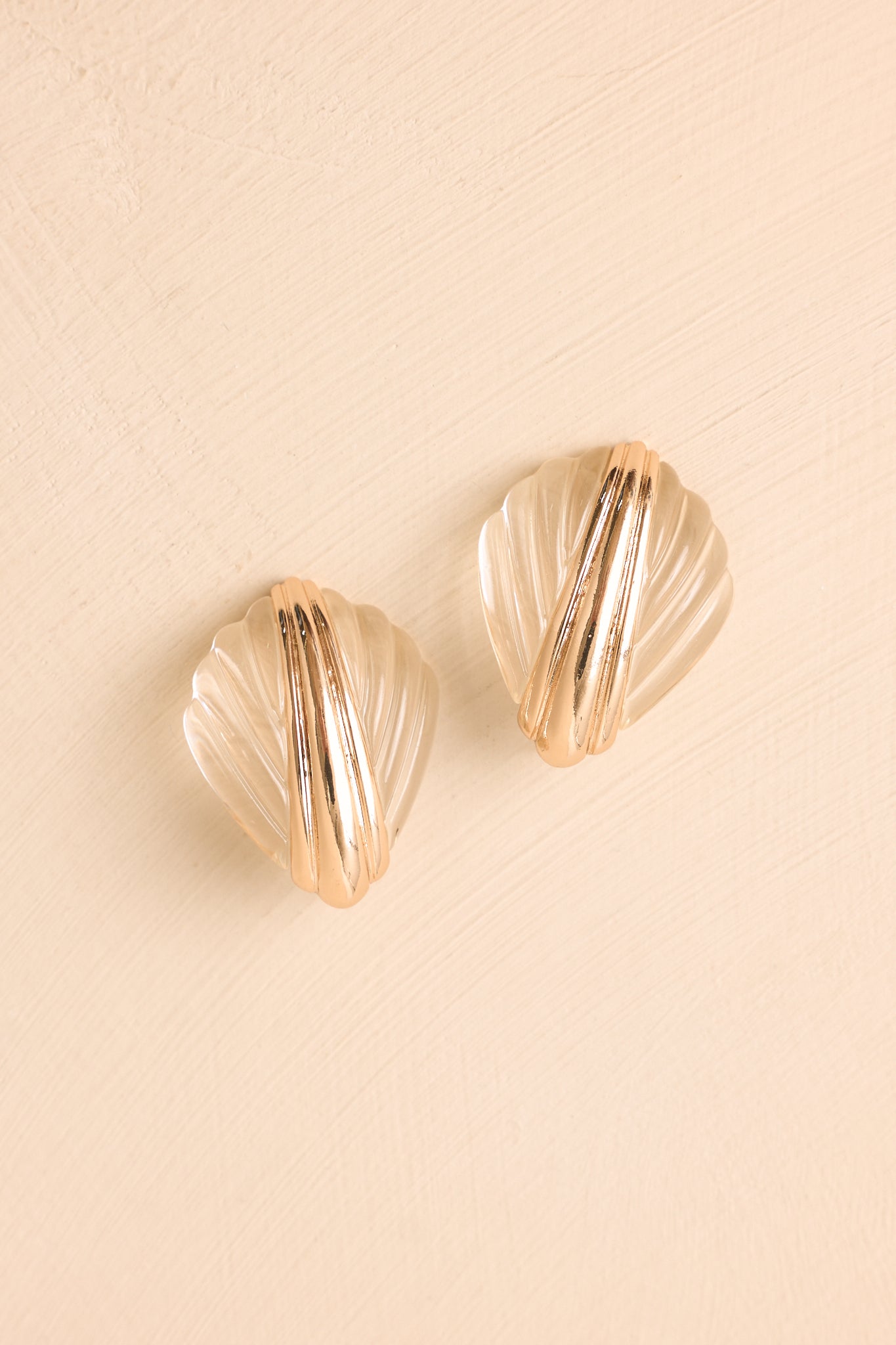 Pair of gold and clear shell earrings displayed on a beige background, featuring acrylic details and polished metallic accents.