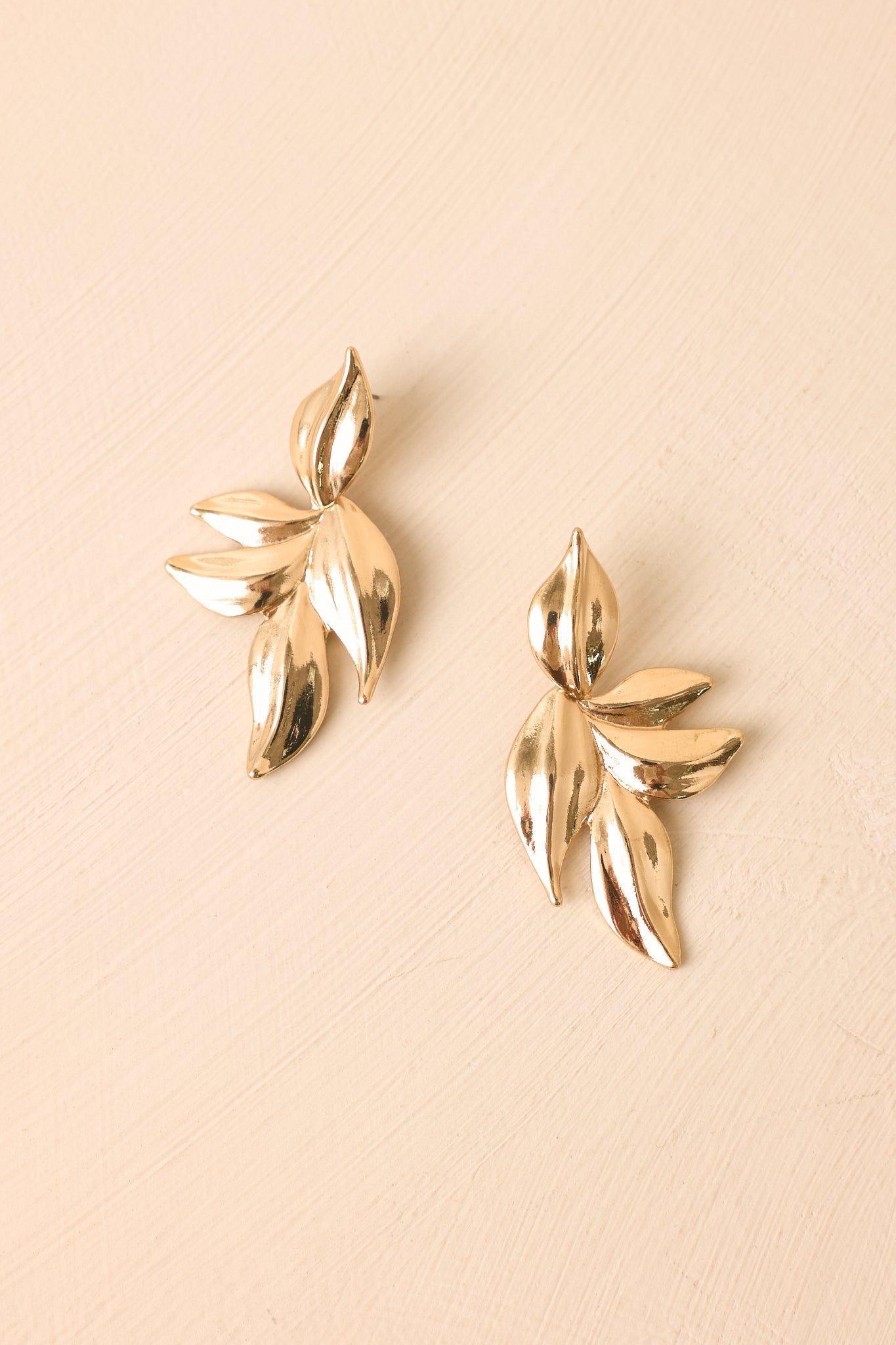 Elegant gold statement earrings on a neutral beige background featuring polished metallic leaves and a bold organic shape.