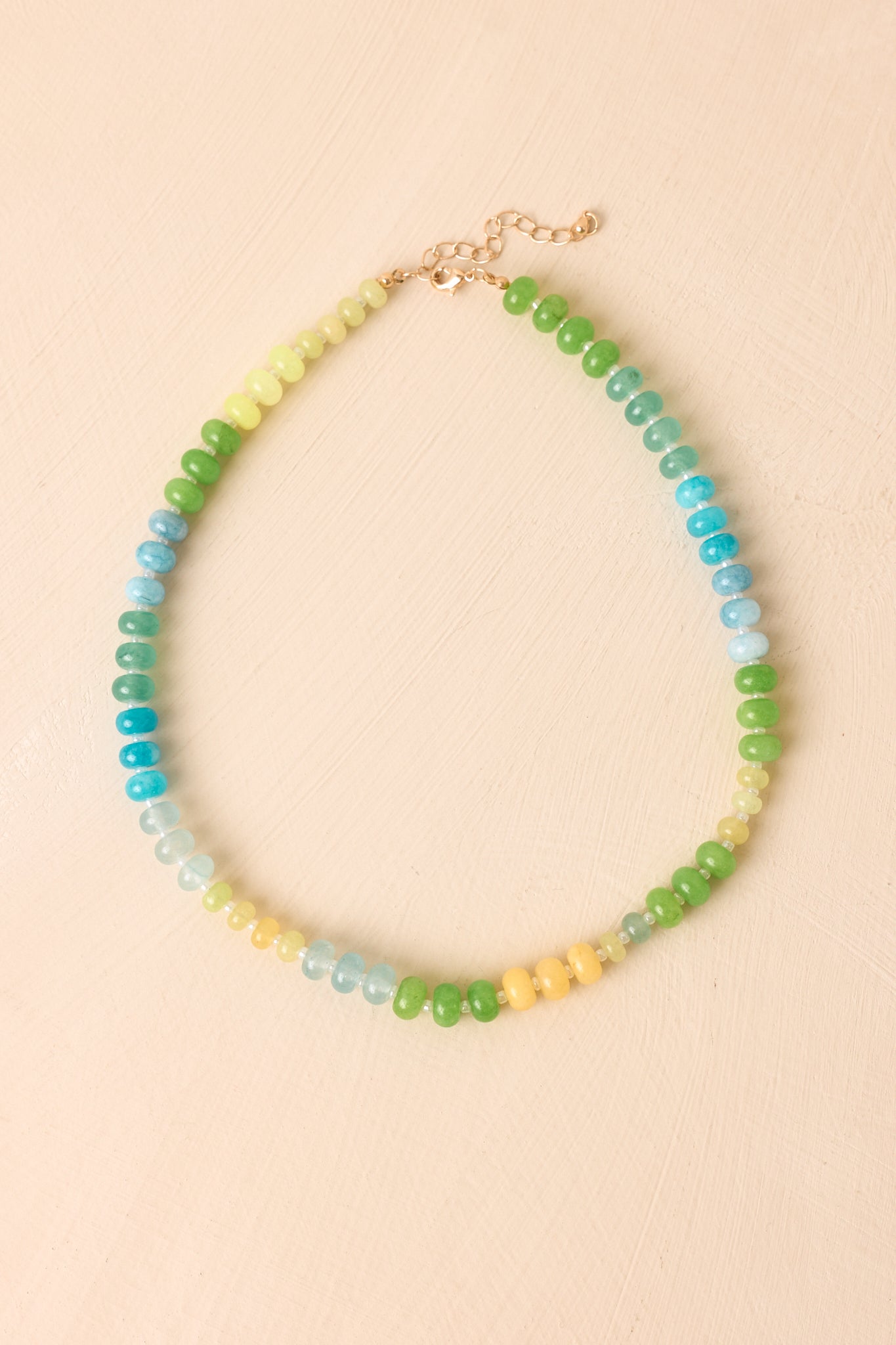 Colorful necklace arranged on a neutral beige background featuring mixed green, aqua, blue, and yellow beads with an adjustable chain extender.