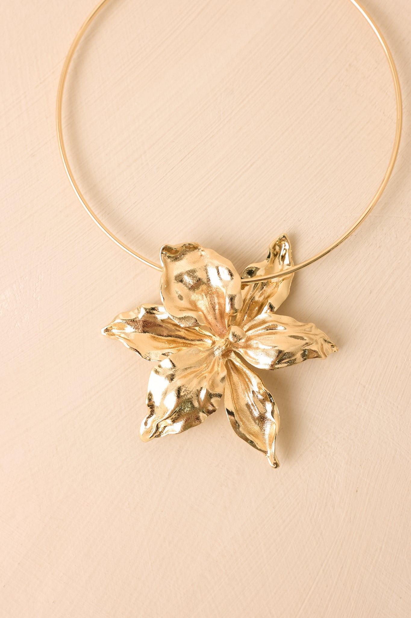 Gold open collar necklace displayed on a beige background, highlighting the oversized floral centerpiece and smooth metallic finish.