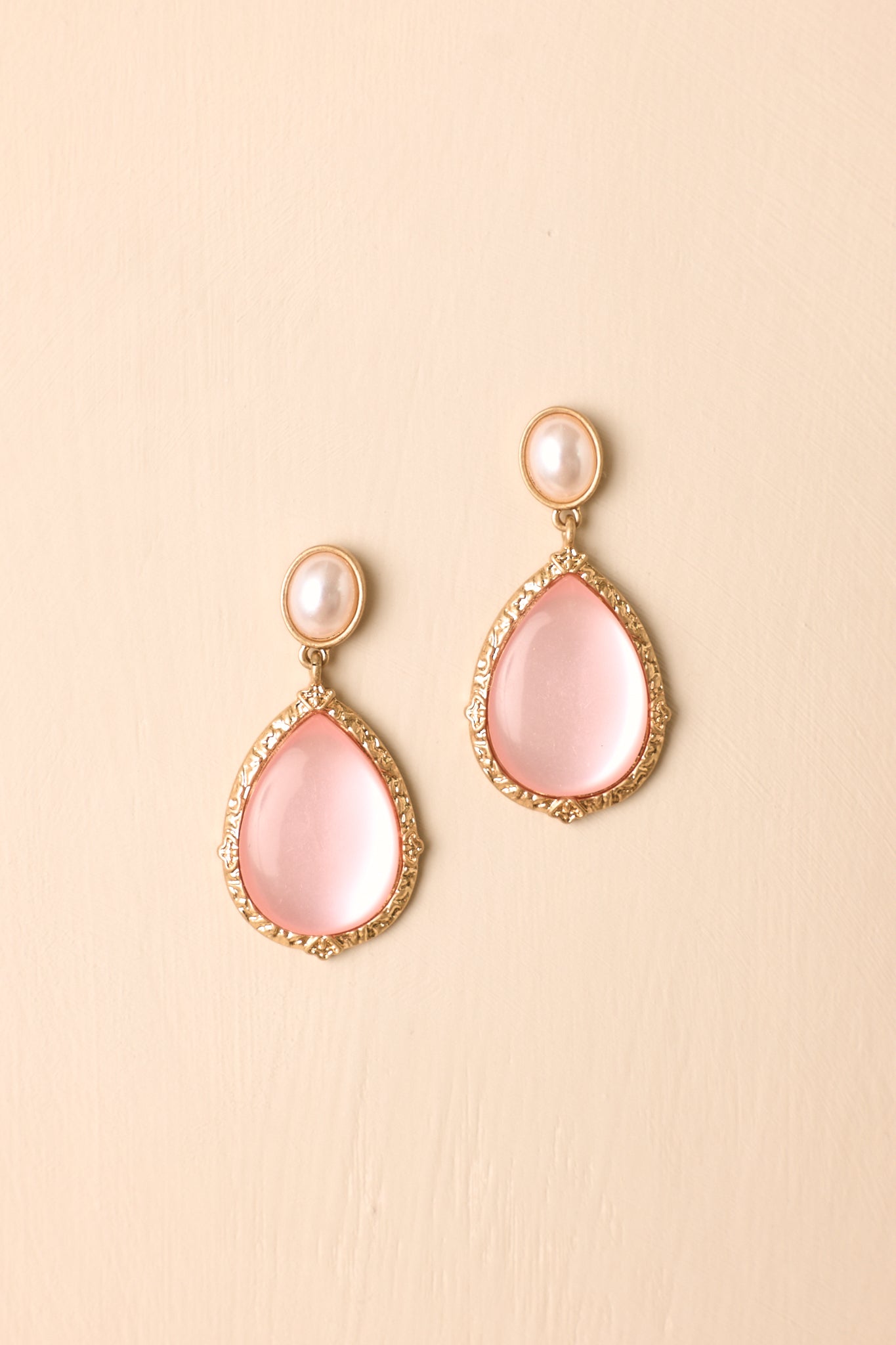 Pair of pink pearlescent earrings displayed on a beige background, featuring teardrop stones and textured gold-tone settings.