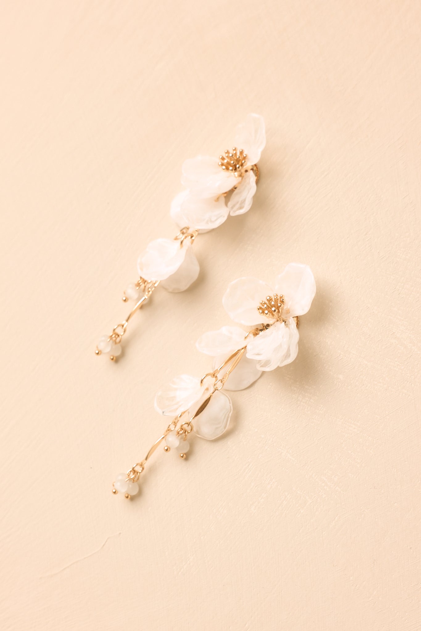 Pair of white floral drop earrings displayed on a beige background, featuring acrylic petals, gold-tone details, and a feminine statement design.