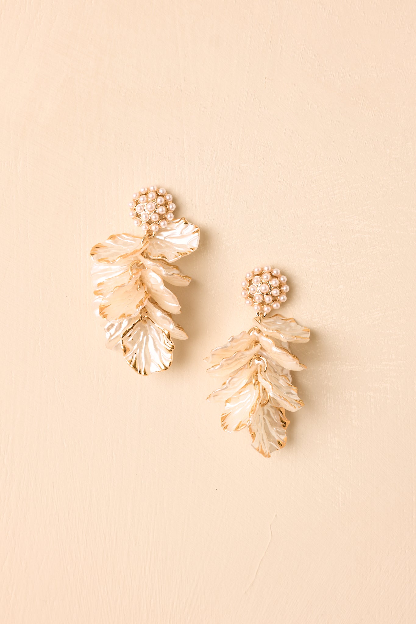 Pair of gold drop earrings displayed on a beige background, featuring pearl cluster tops and layered ivory petal charms.
