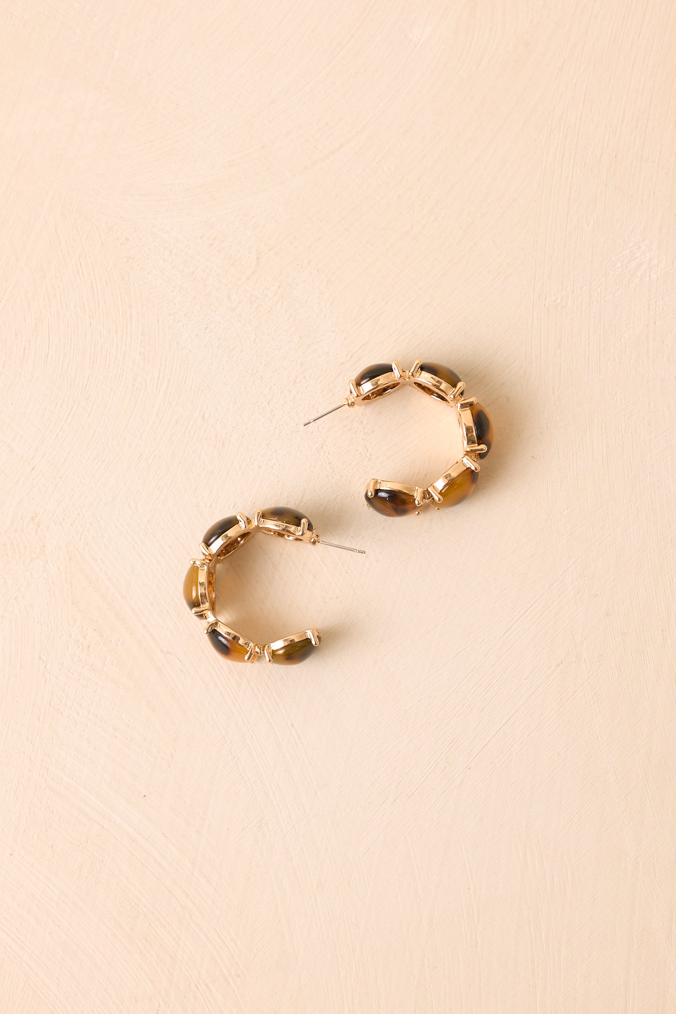 Open hoop earrings with a classic tortoise pattern and gold-tone frame, styled on a neutral beige background for a refined presentation.