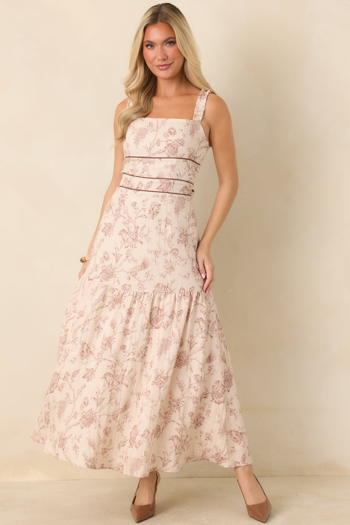 A woman modeling a sleeveless tiered maxi dress with a dusty pink floral toile pattern on a cream background, paired with brown heels, set against a plain beige studio backdrop.