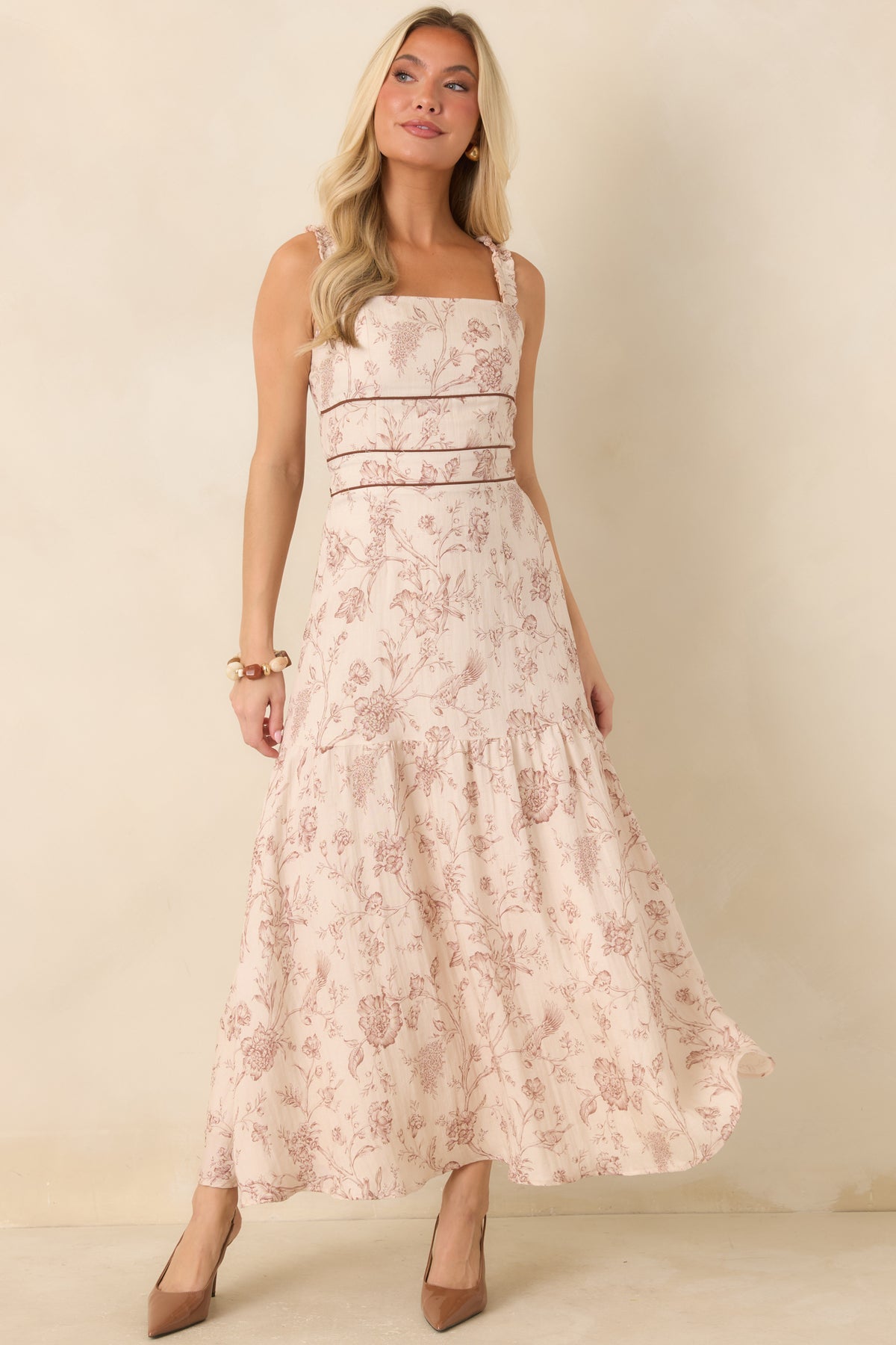A woman modeling a sleeveless, tiered maxi dress with a light pink and beige floral pattern and thin brown shoulder straps, standing against a plain beige studio backdrop.