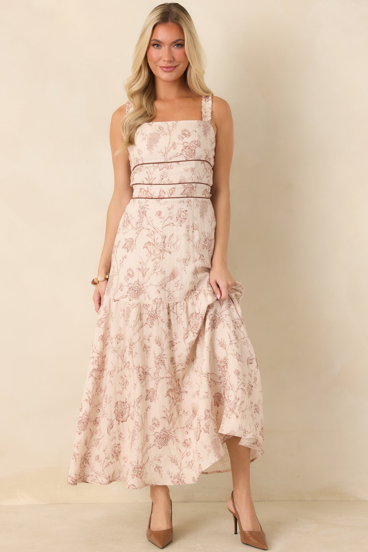 A woman modeling a sleeveless, tiered maxi dress with a dusty rose floral pattern on a cream background, standing against a plain beige studio backdrop.