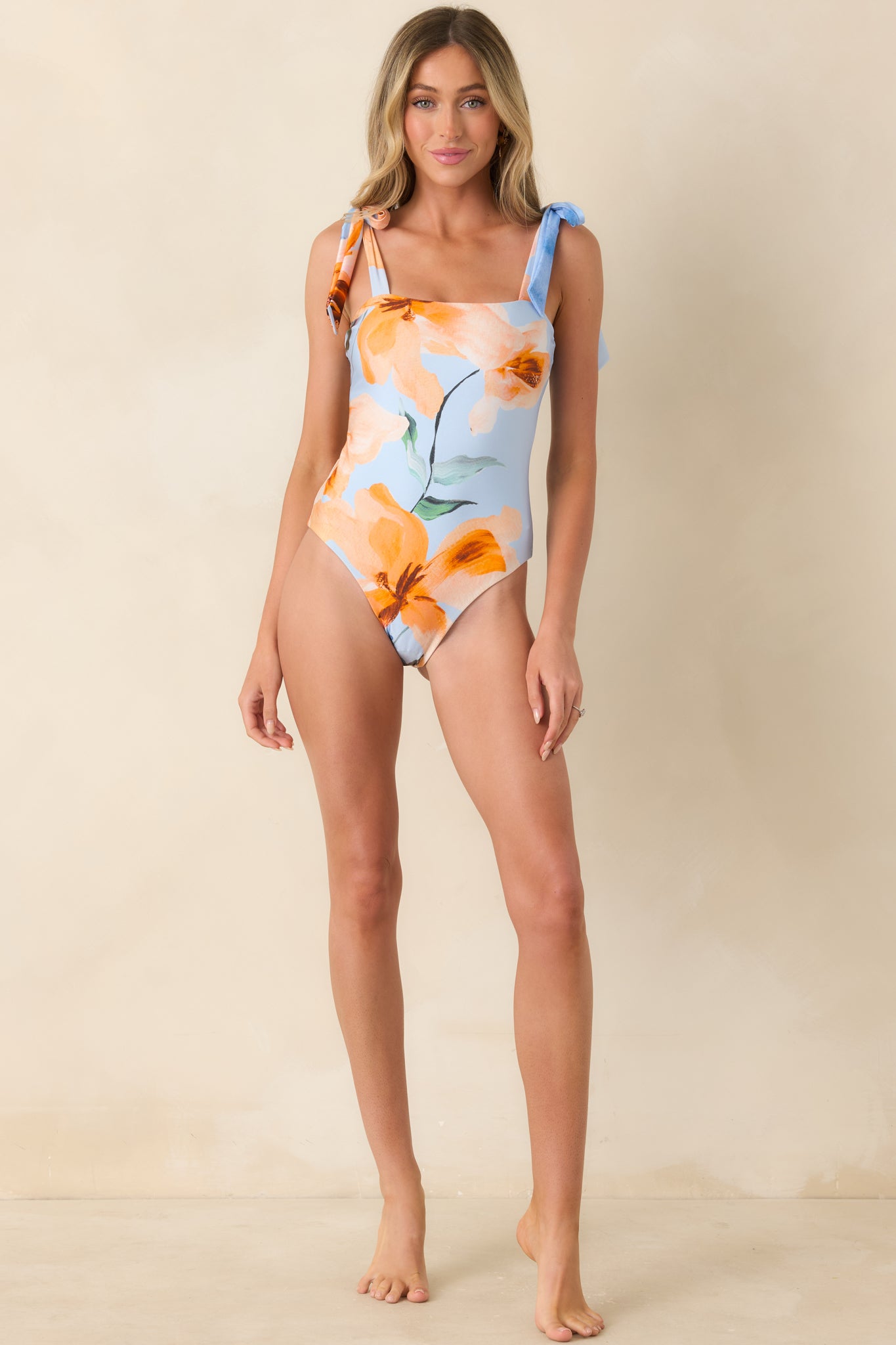Blue and gold floral one-piece featuring an adjustable tie-strap design, clean neckline, and modern fitted silhouette with smooth lines.