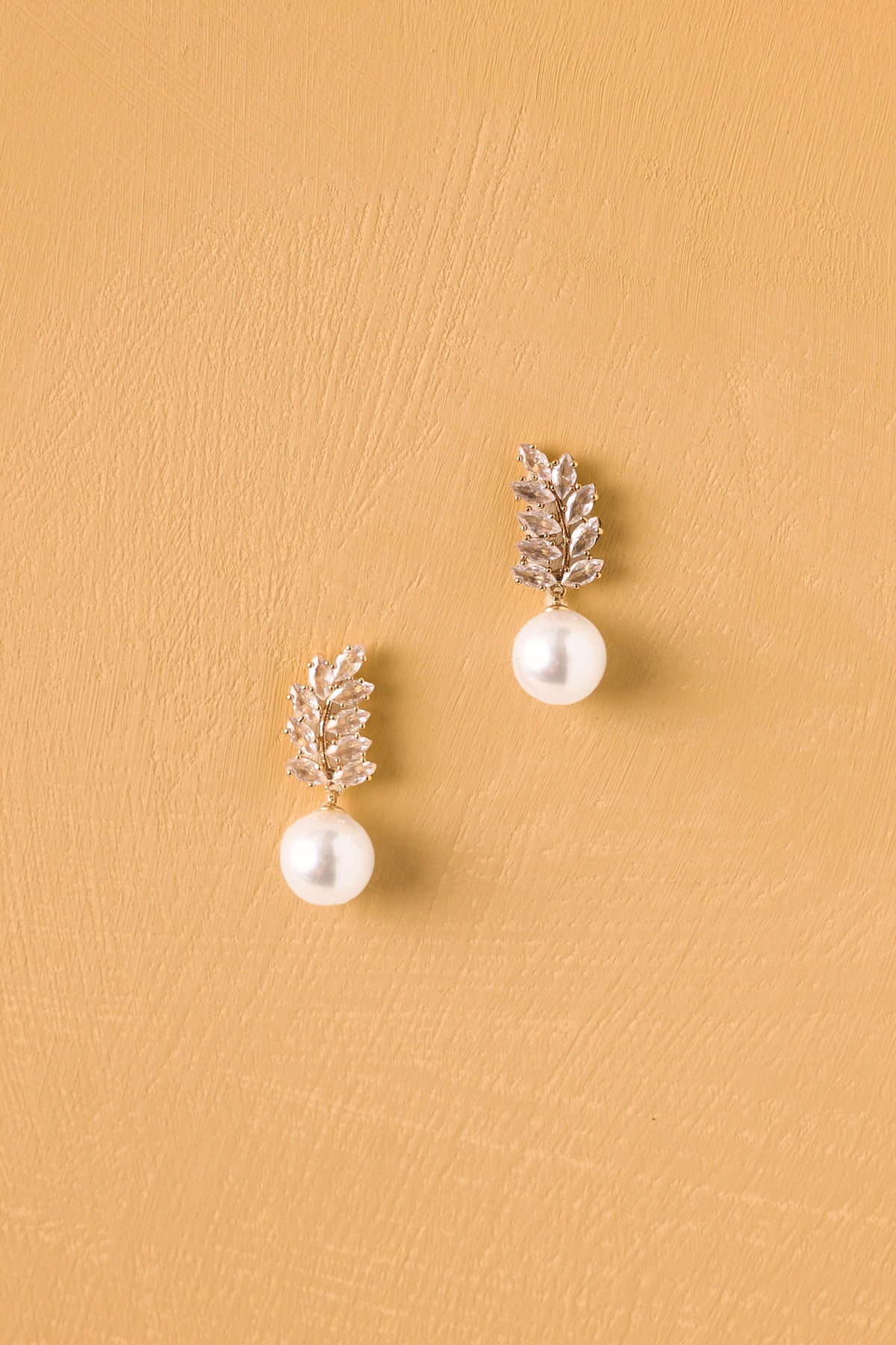 Pearl drop earrings featuring a gold leaf design embellished with clear crystals in front of a golden yellow backdrop. 