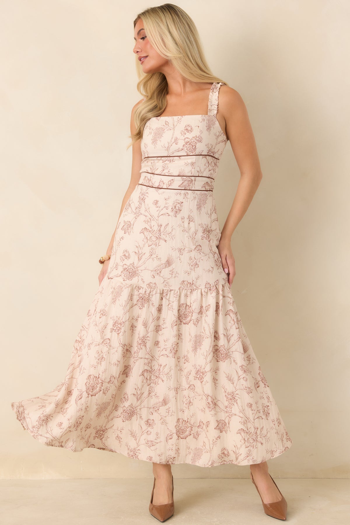 A woman modeling a sleeveless, tiered maxi dress with a dusty pink floral pattern on a cream background, set against a plain beige studio backdrop.