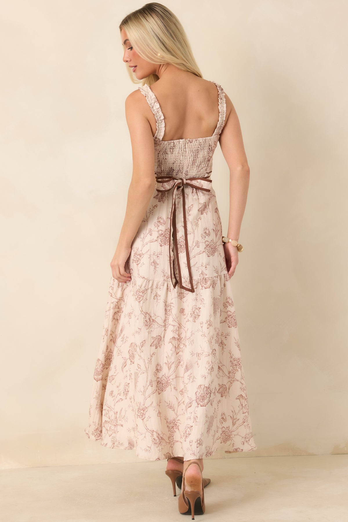 A woman wearing a sleeveless maxi dress with a floral toile pattern in dusty pink and beige, featuring lace straps, a tie waist, and brown high heels, shown from the back against a warm beige studio backdrop.