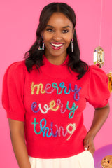 Front view of this sweater that features a slight turtle-neck, short puff sleeves with ribbed cuffs, a ribbed hem, and multi-colored tinsel script. 