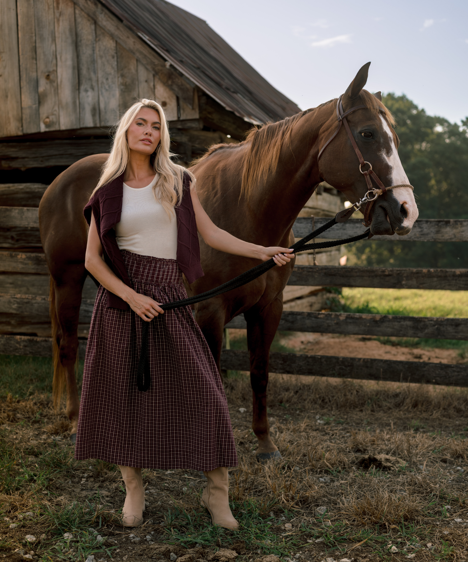 A woman stands outdoors in front of a rustic wooden barn and fence, holding a brown horse on a lead rope. She is wearing a white tank top, a dark burgundy sweater around her shoulders, a dark burgundy plaid midi skirt, and beige ankle boots.