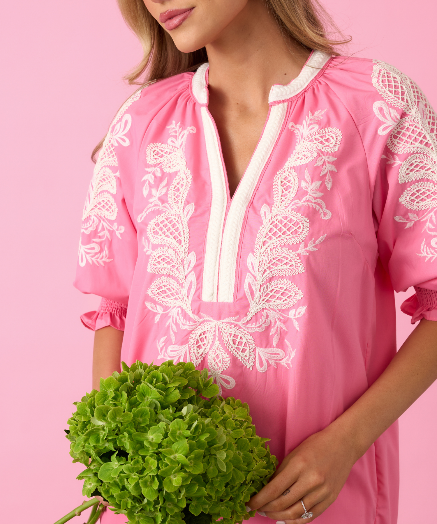 Woman wearing a pink embroidered dress holding green hydrangeas against a pink background.