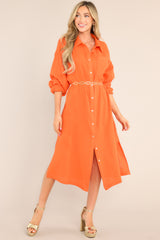 This orange dress features a collared neckline, functional buttons down the front, a front pocket on the left side of the bust, long sleeves with a cuff secured by a functional button, and two slits up the bottom hemline ending just below the knee.