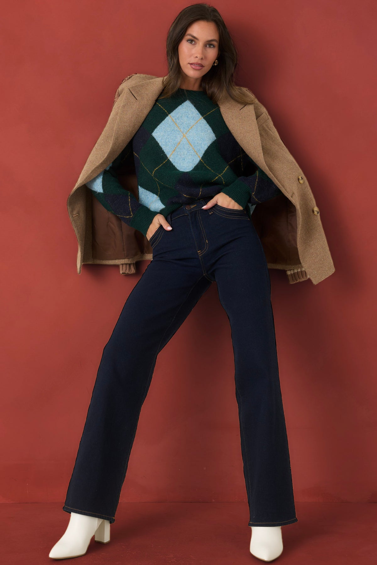 A woman models a stylish outfit consisting of a green and light blue argyle sweater, dark blue jeans, a tan coat, and white heeled boots, posed against a solid terracotta red background.