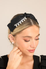 Top view of this headband that features a cloth covered metal piece, a black bow attached to the top, a thin body, diamond & pearl detailing, and rubber tips.