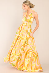 This yellow and orange dress features a halter neckline with an adjustable self-tie at the back of the neck, an open back, an elastic waistband, two functional pockets, a long flowy skirt, and a vibrant pattern throughout.