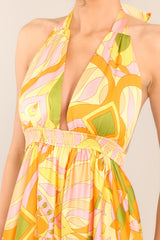 Close up view of this dress that features a halter neckline with an adjustable self-tie at the back of the neck, an open back, an elastic waistband, two functional pockets, a long flowy skirt, and a vibrant pattern throughout.