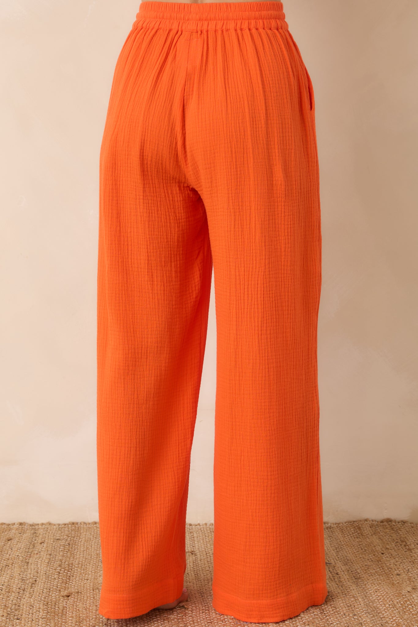 Back view of vibrant orange wide-leg pants featuring a relaxed fit and elastic waistband in lightweight textured cotton.