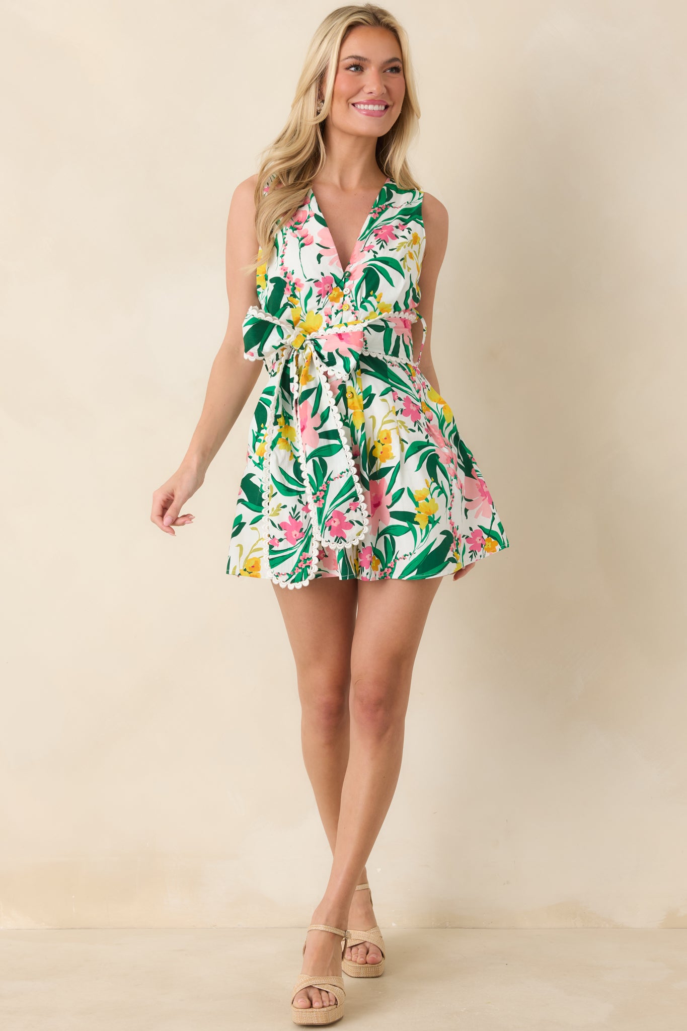 Tropical floral romper in shades of green, pink, and yellow, featuring a sleeveless V-neckline, button-front bodice, scalloped trim, and coordinating tie belt above flared shorts.