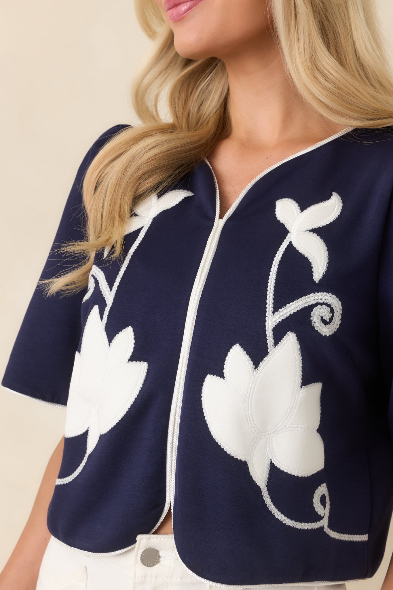 Close-up highlighting the ivory floral appliqué, vine embroidery, and contrast piping along the neckline and sleeves.