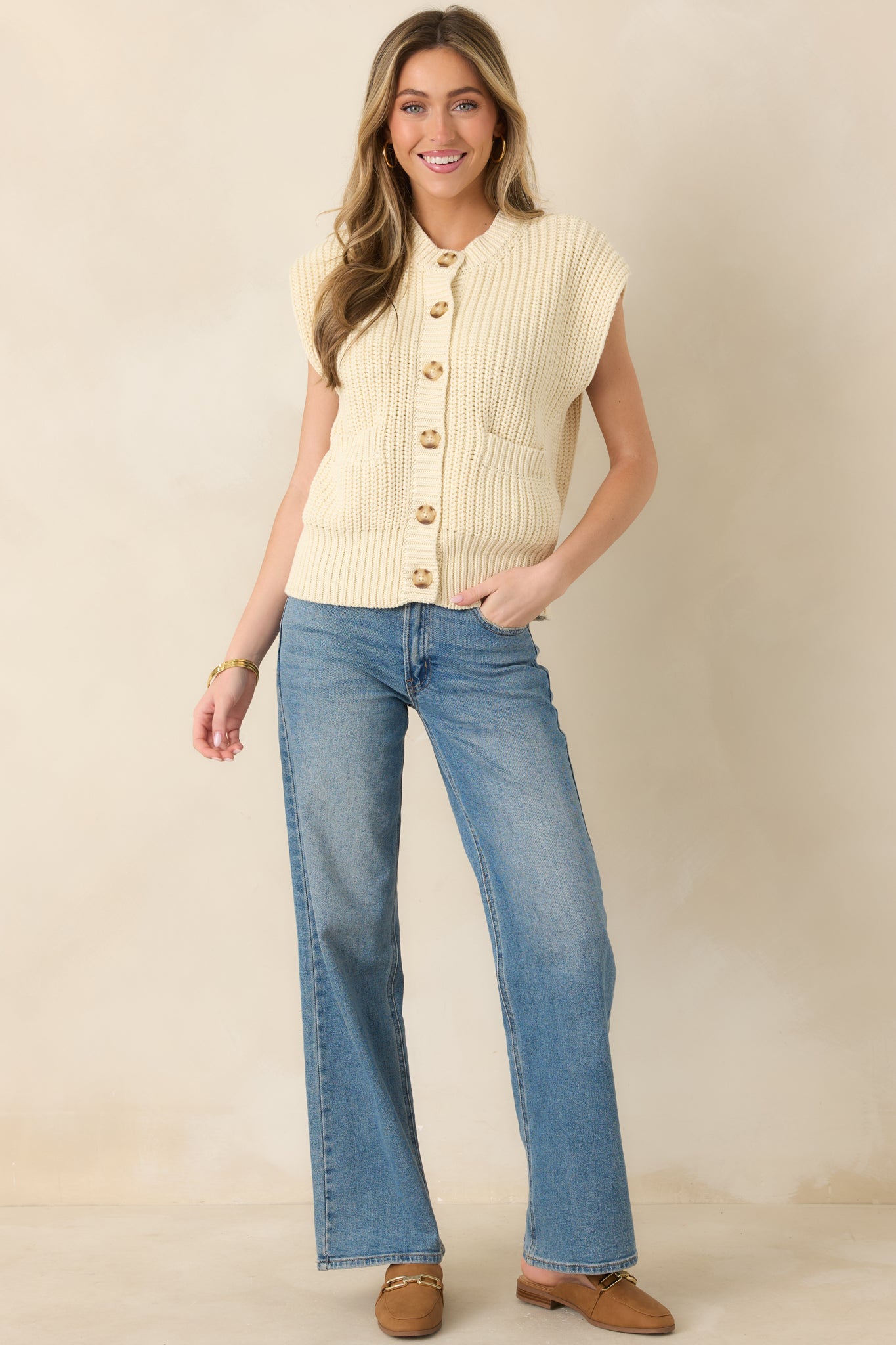 Front view of an ivory sleeveless knit cardigan with button-front closure, ribbed trim, and front patch pockets.
