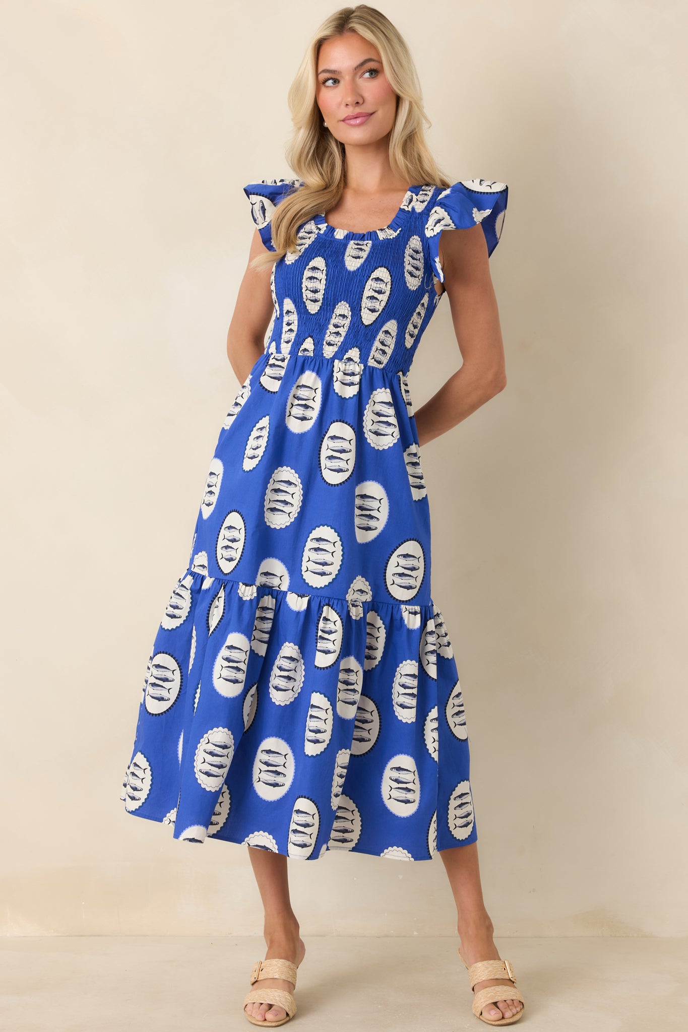 Royal blue tiered dress in breathable cotton with scattered fish print, detailed with flutter sleeves and a flexible smocked bodice for a comfortable, flattering shape.