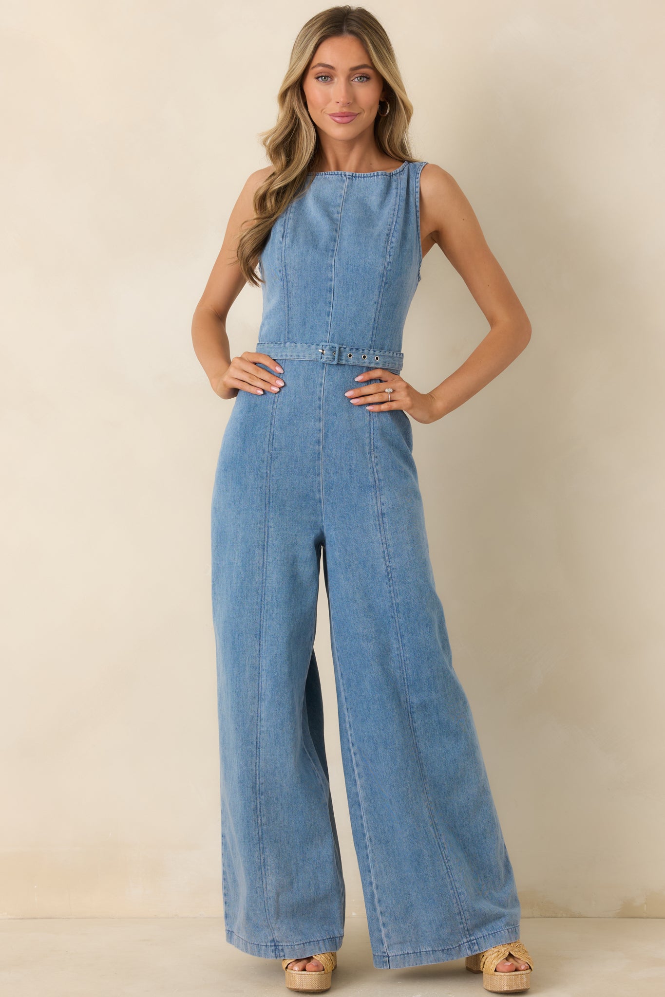 Light wash denim jumpsuit with sleeveless design, high neckline, and fitted waist with subtle belt detail