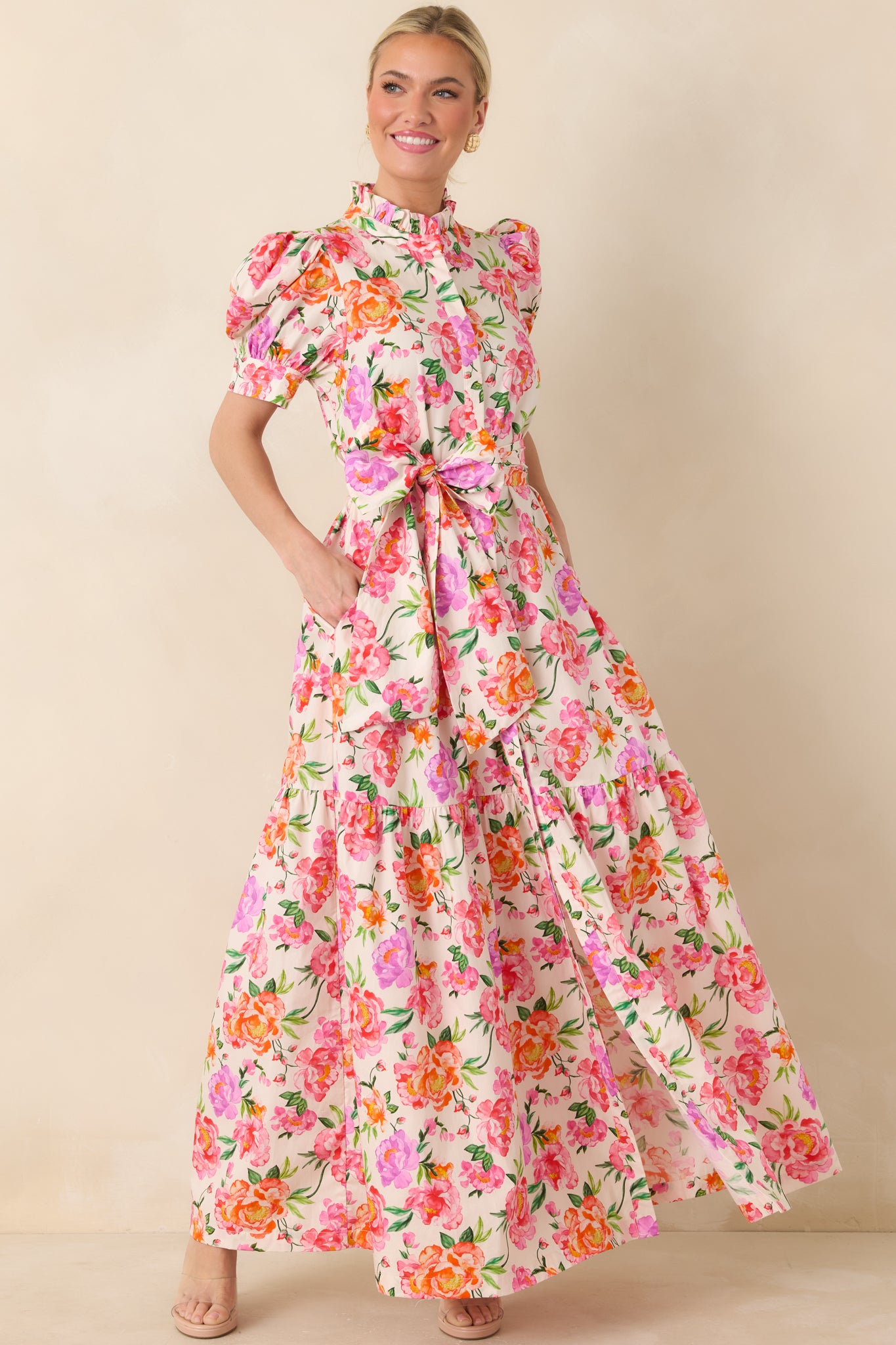 Pink multi floral maxi dress with ruffled high neckline, softly puffed sleeves, adjustable waist tie, and a flowing tiered skirt.