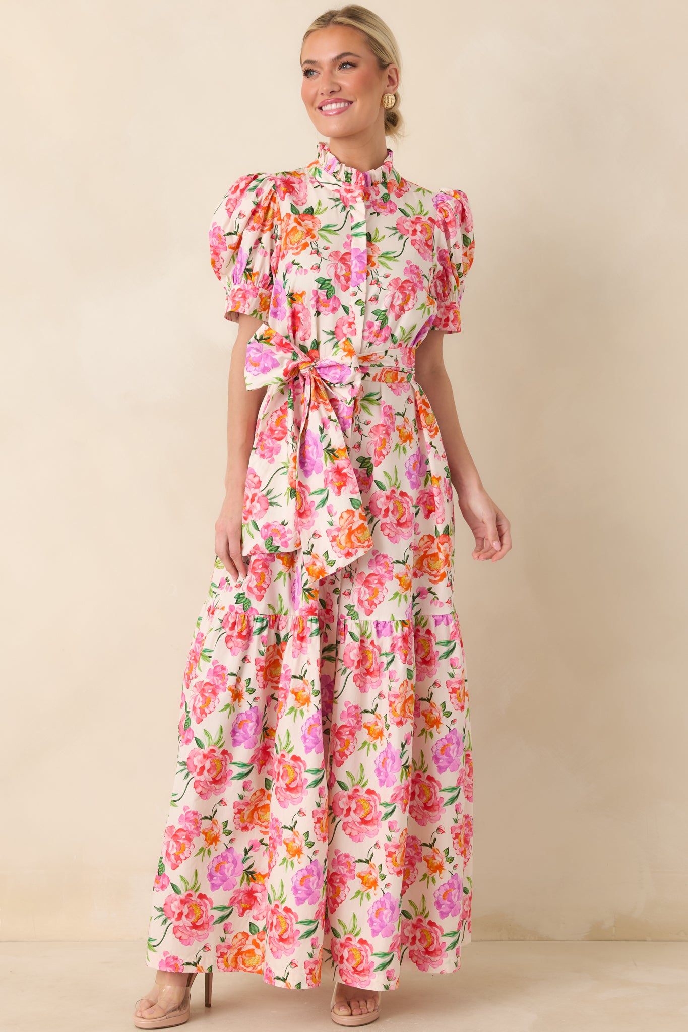 Feminine pink floral maxi dress with ruffled neckline, soft puff sleeves, waist-defining tie, and tiered skirt for graceful movement.