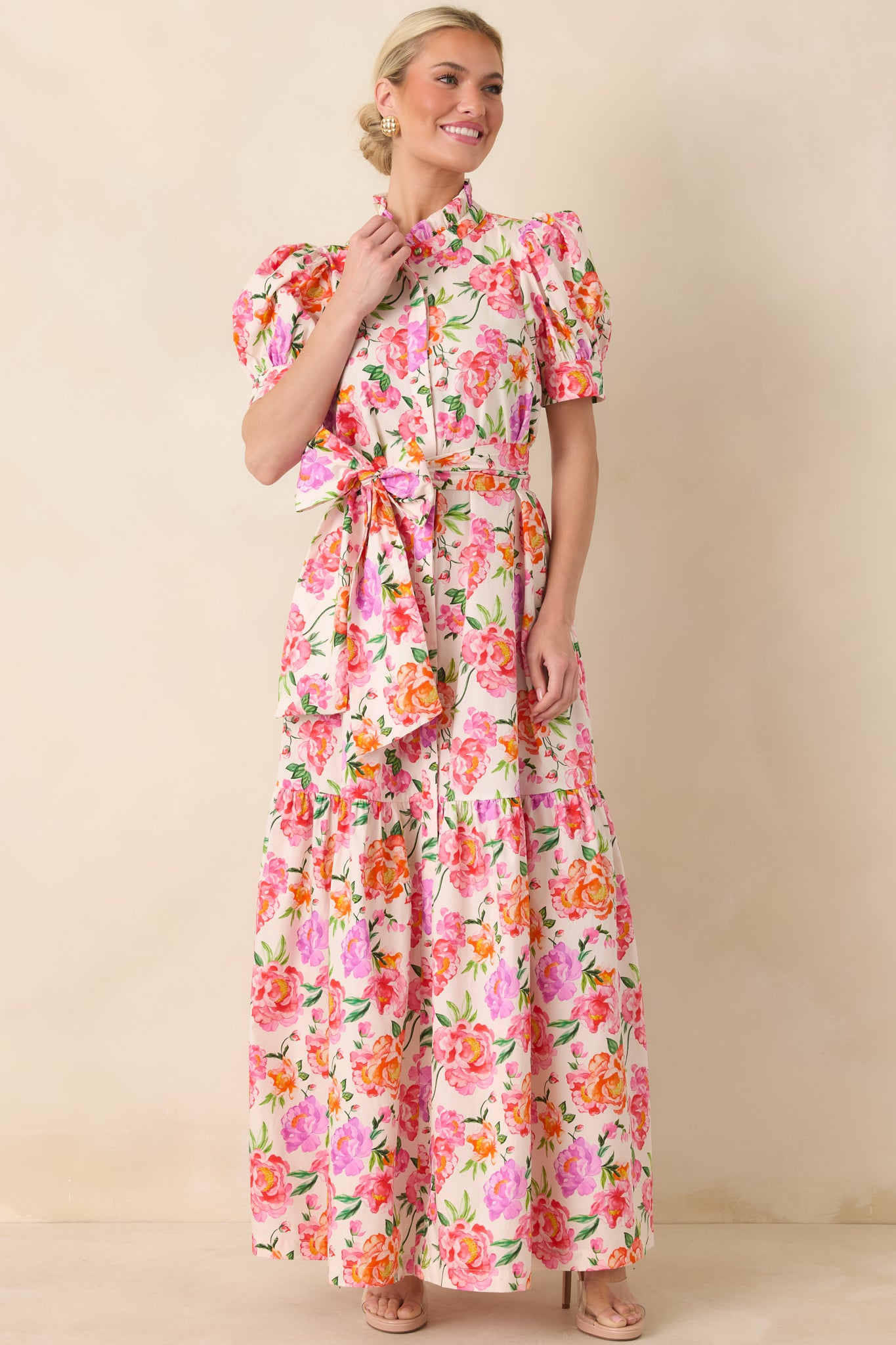 Tiered maxi dress in pink multi floral cotton with puffed sleeves, ruffled high neckline, and adjustable tie at the waist.