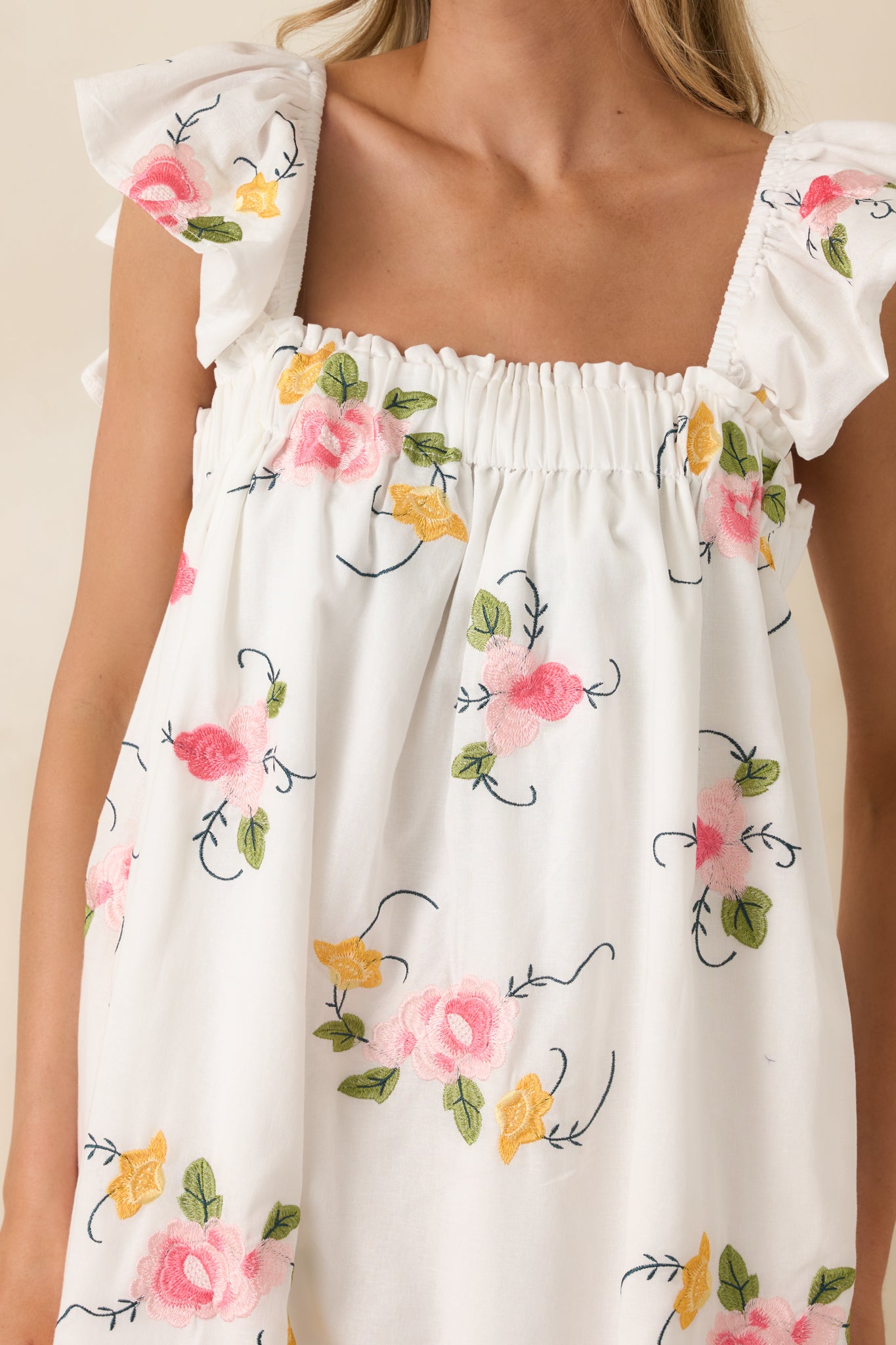 Detailed view of the pink and golden floral embroidery against the white cotton linen fabric, highlighting the textured stitching and ruffled strap detail.