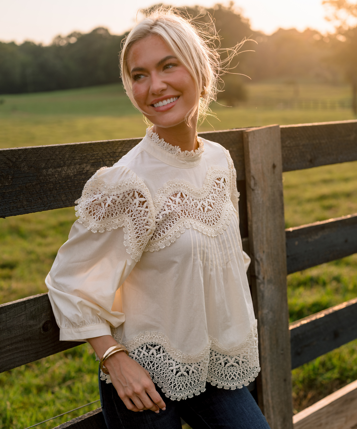 A smiling blonde woman leans against a rustic wooden fence in a grassy field at sunset, wearing a cream lace-trimmed blouse with puff sleeves and dark blue jeans.