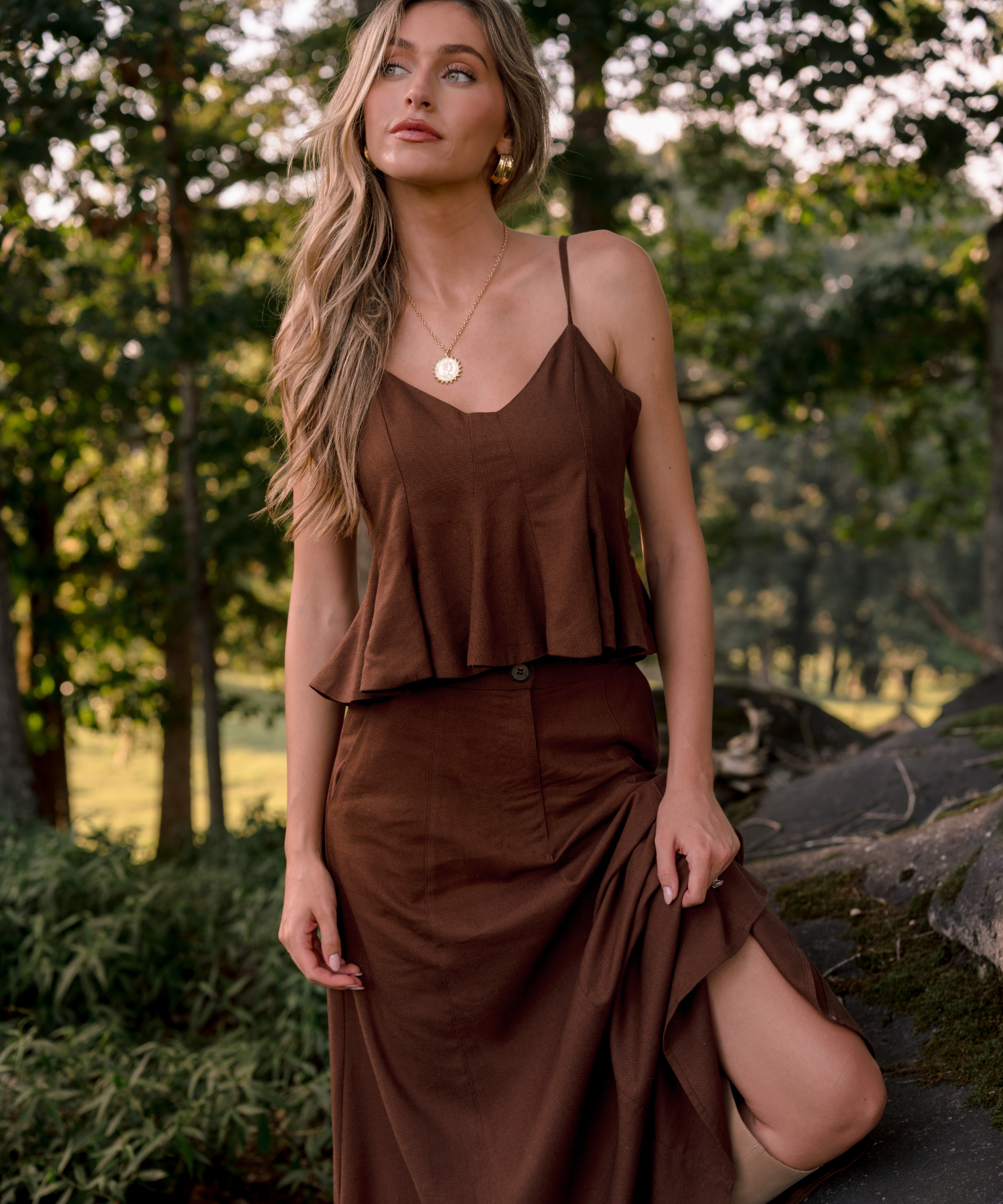A woman wearing a brown two-piece set consisting of a sleeveless peplum top and a matching high-waisted skirt, posing outdoors in a wooded setting.
