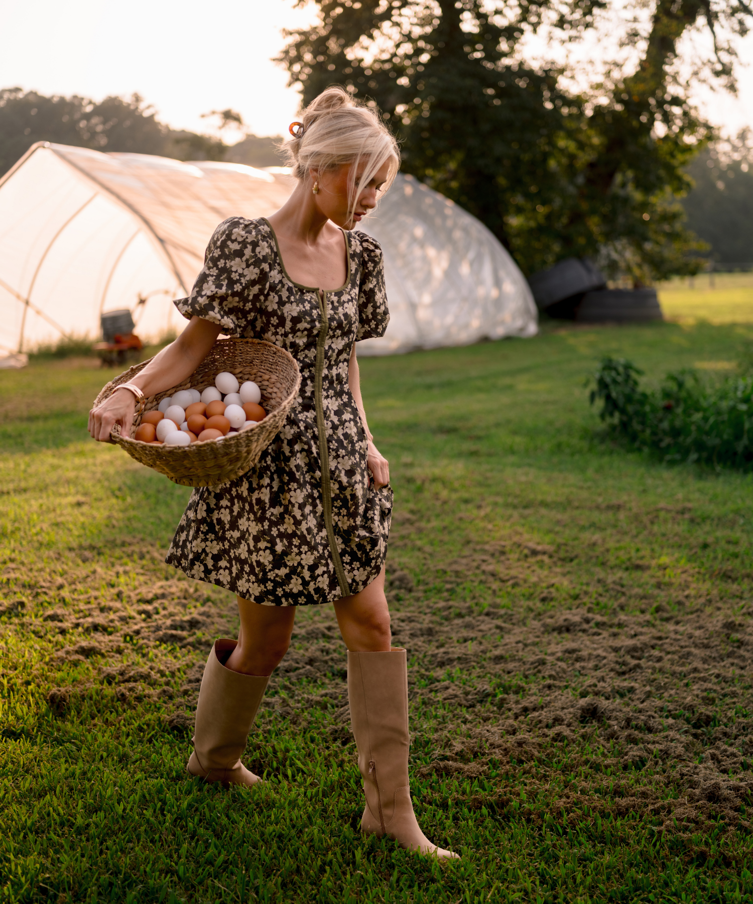 A woman in a short floral dress and beige boots walks across a grassy field carrying a basket of eggs, with a greenhouse and trees in the background.