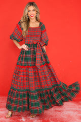 Full body view of this dress that features a square neckline, puff sleeves with elastic cuffs, a fully smocked bust, a self-tie belt at the waist, and a long, exaggerated skirt with alternating plaid patterns. 