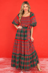 This red dress features a square neckline, puff sleeves with elastic cuffs, a fully smocked bust, a self-tie belt at the waist, and a long, exaggerated skirt with alternating plaid patterns. 