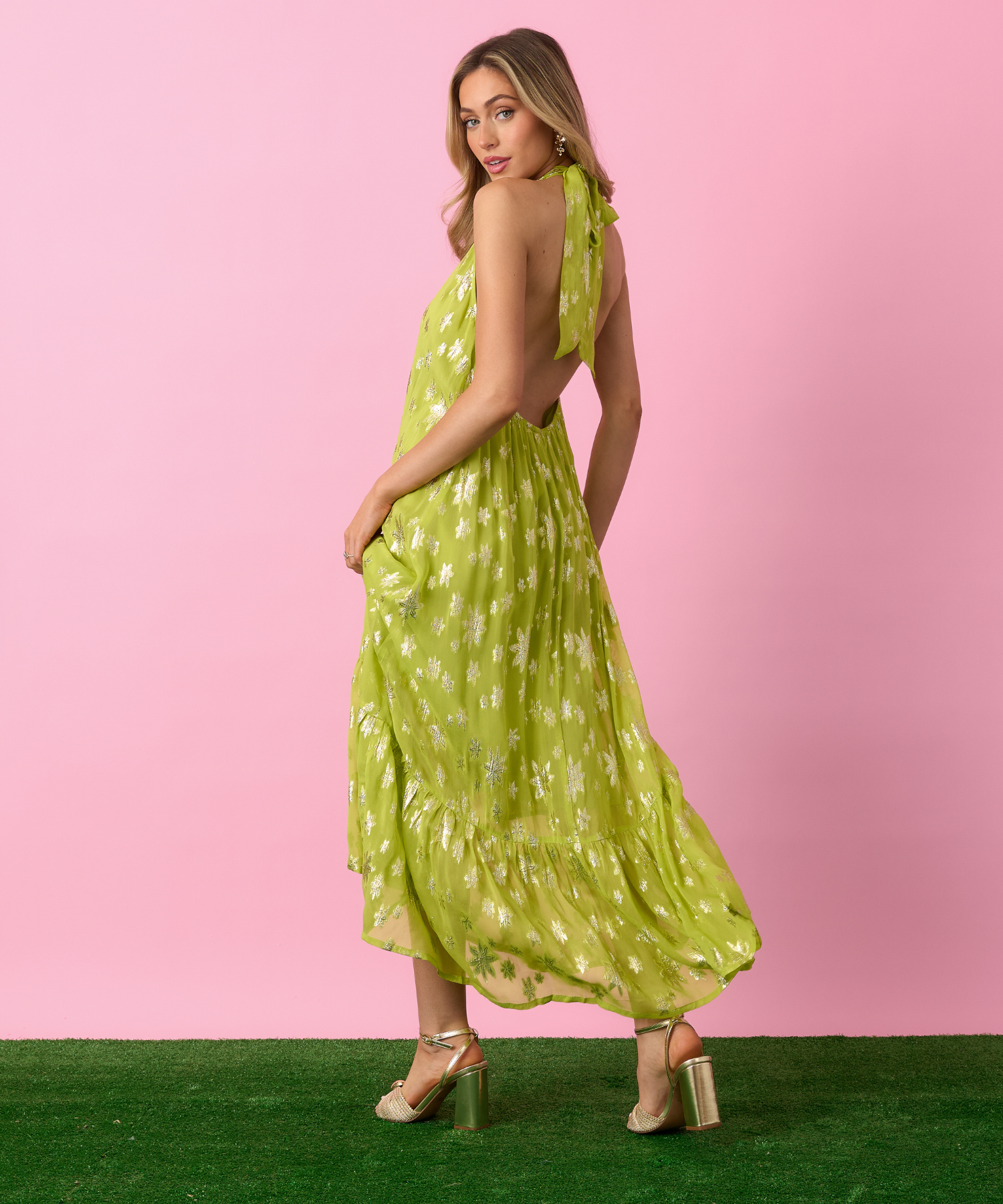 Woman in a green dress with metallic floral patterns standing against a pink background.