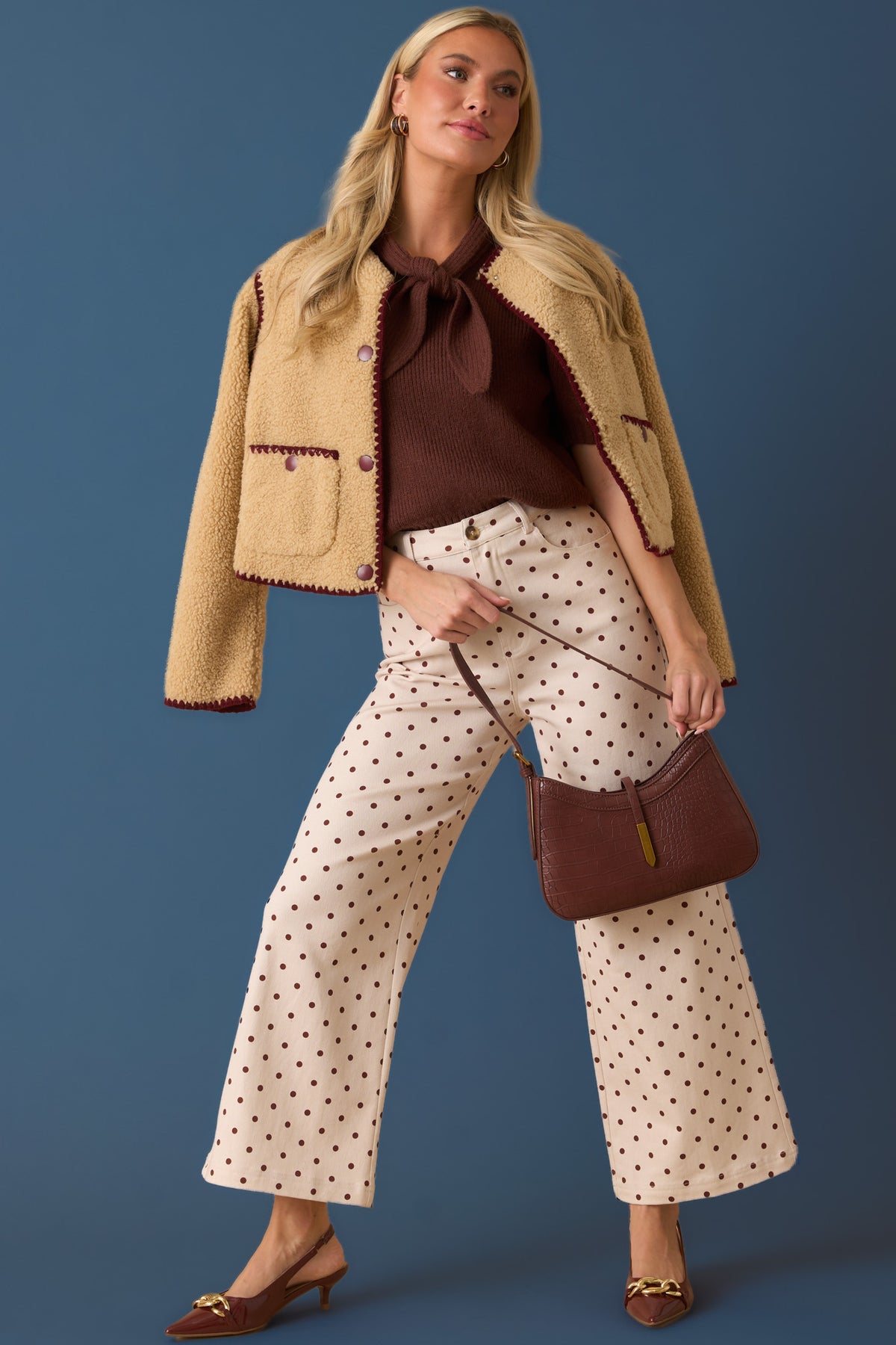 A woman models a stylish outfit featuring a beige textured jacket with dark brown trim, a dark brown top with a large bow, cream wide-leg pants with brown polka dots, and brown pointed-toe flats, while holding a matching brown handbag against a solid blue background.