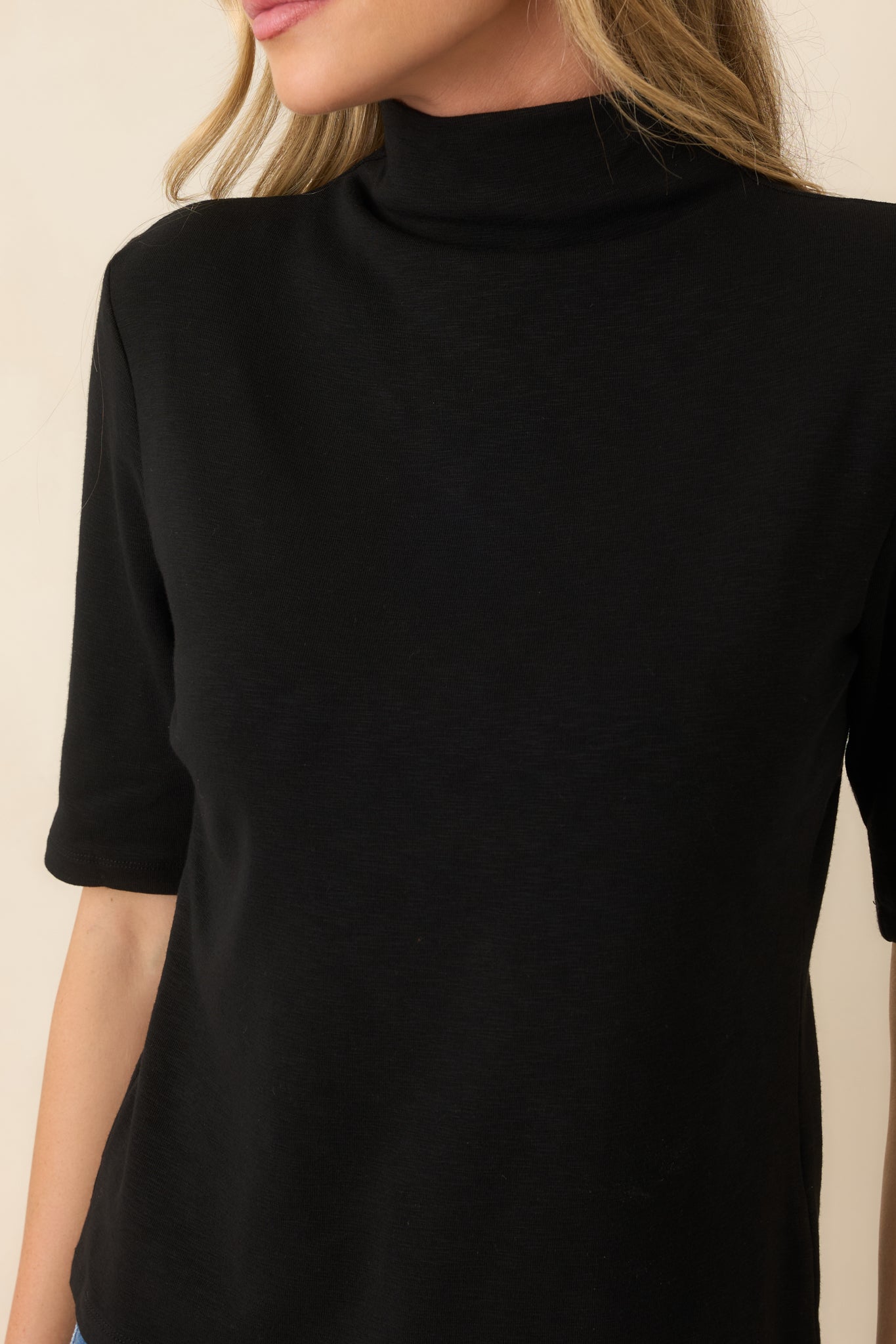 A close-up of a woman wearing a black short-sleeve mock neck top against a light beige background.