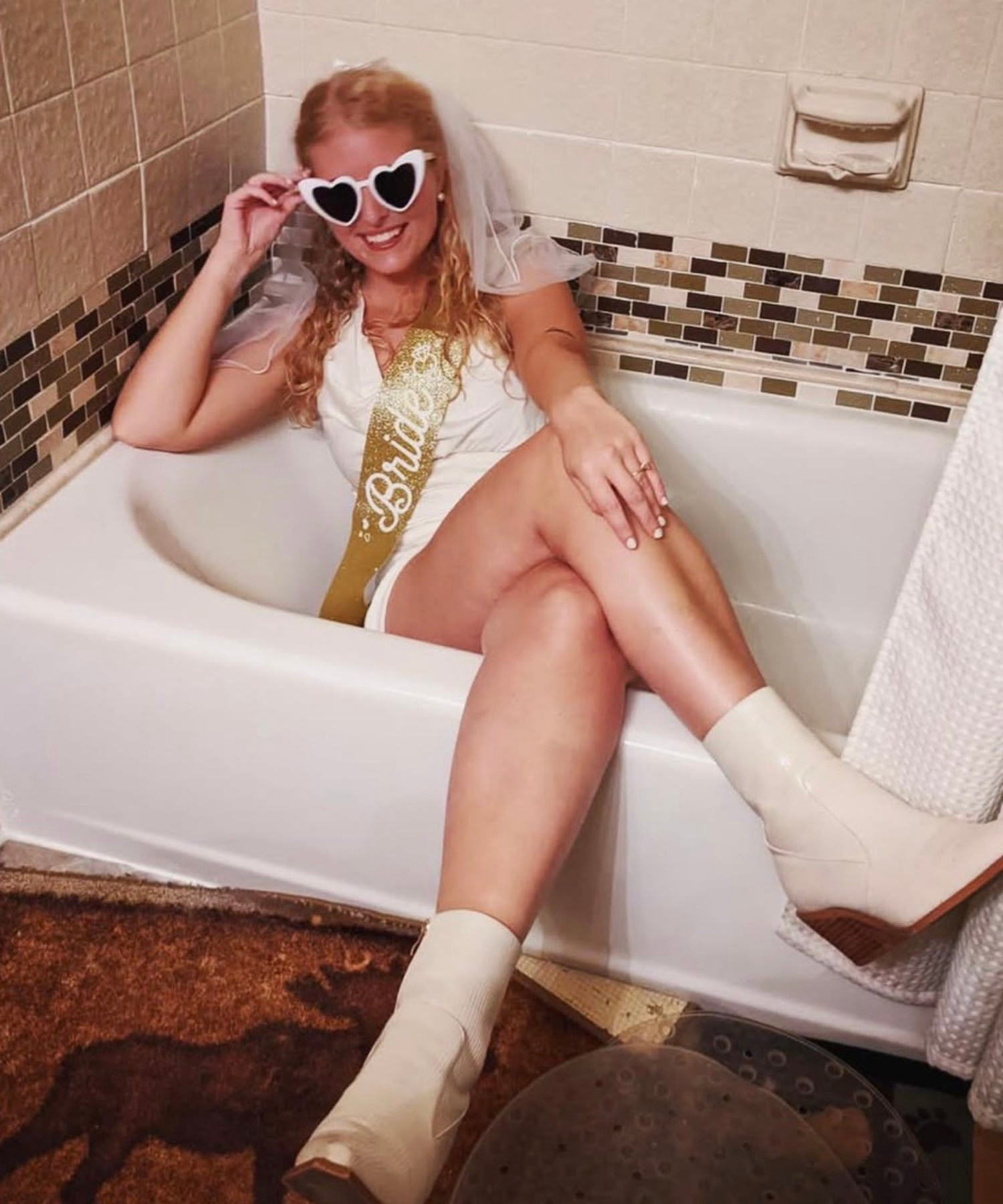 Woman in a bathtub wearing a 'Bride' sash and heart-shaped sunglasses.