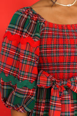 Close up view of this dress that features a square neckline, puff sleeves with elastic cuffs, a fully smocked bust, a self-tie belt at the waist, and alternating green and red plaid patterns. 