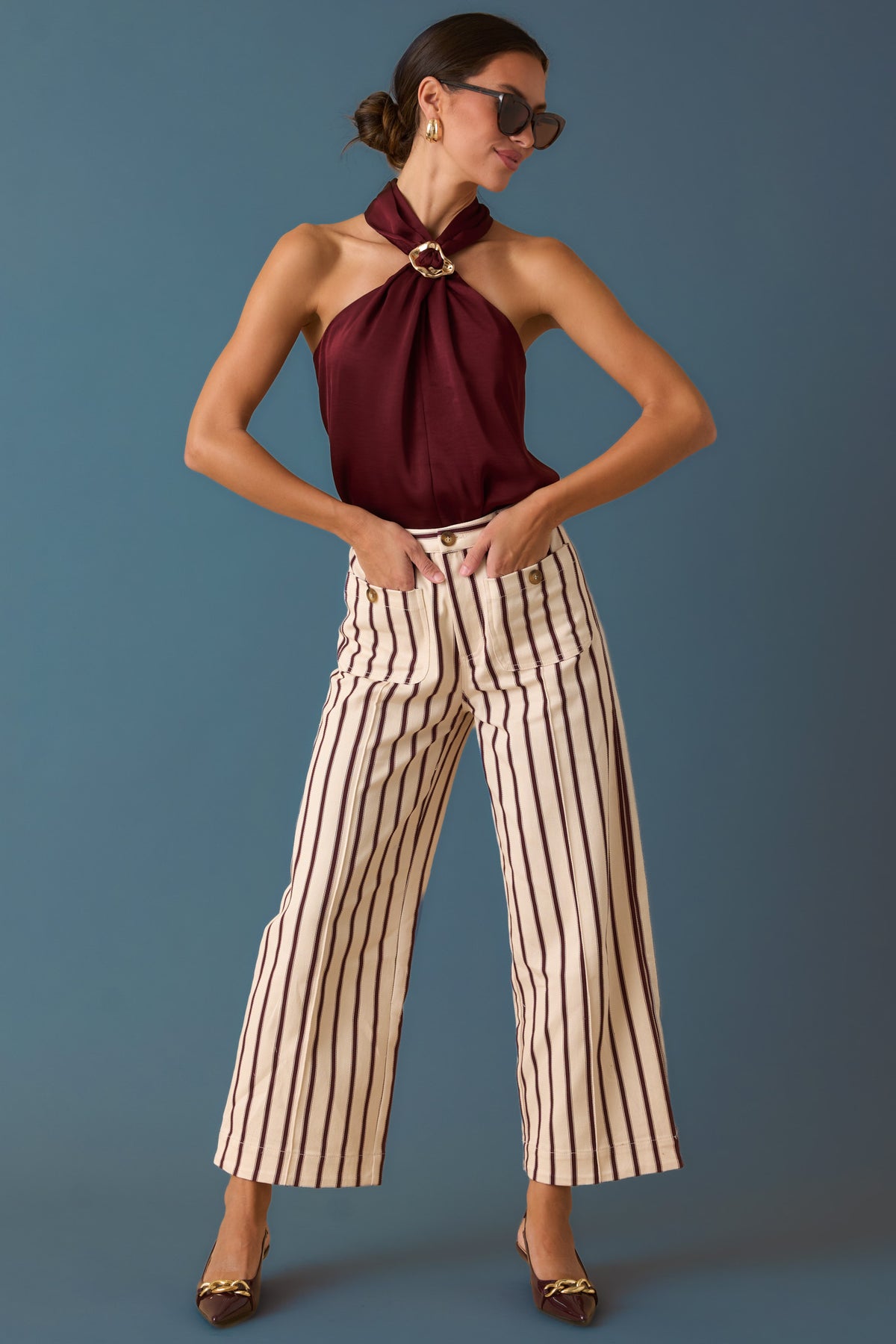 Woman in a burgundy stain halter top and striped pants against a blue backdrop. 
