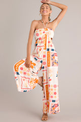 Front full body view of a patterned jumpsuit featuring a halter tassel tie neckline, an elastic waist, cutouts at the waist, functional pockets, and a smocked insert in the back of the bust.